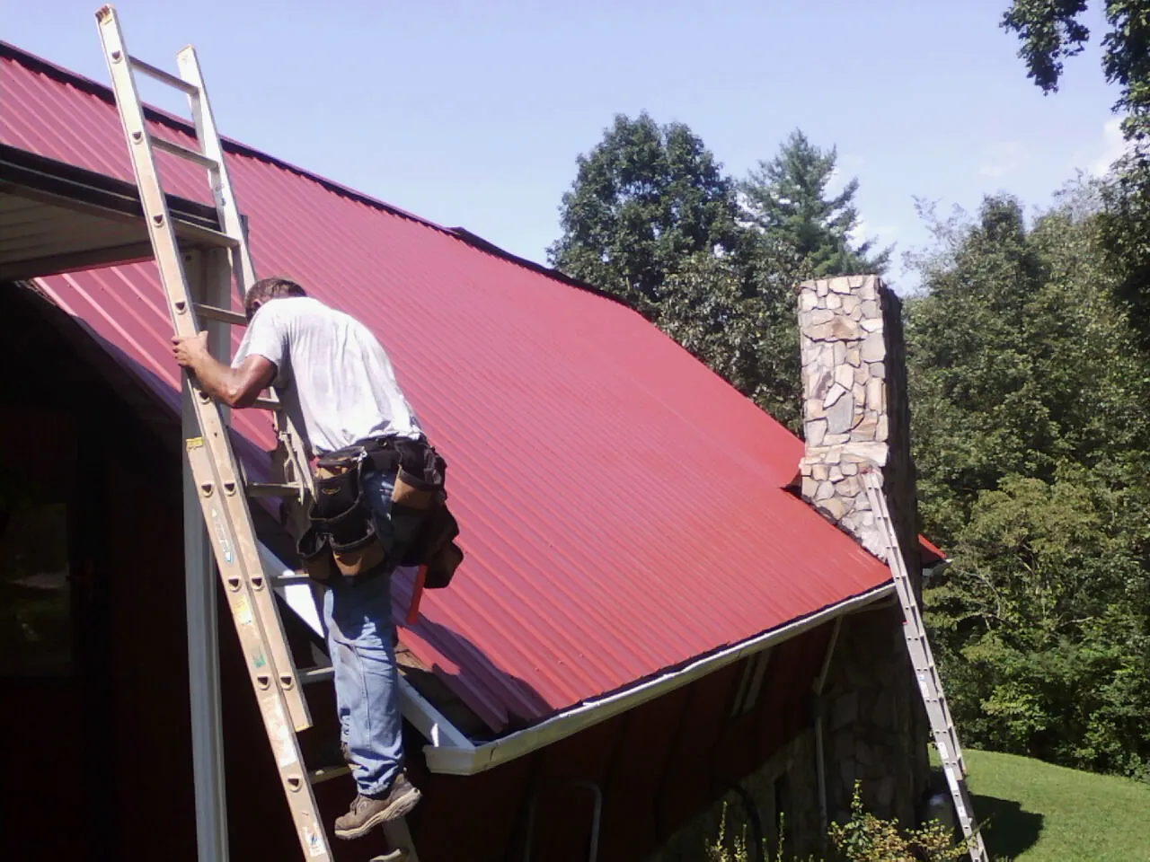 Roofer on a ladder beside a red metal roof and stone chimney — Soffit & Fascia Repair by Roofing Brothers in Verbena